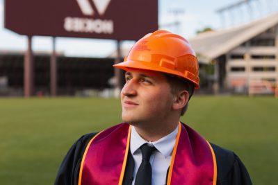 Alex Sherbak, stands on Virginia Tech practice field in an orange hardhat and graduation garb, preparing for career in construction.